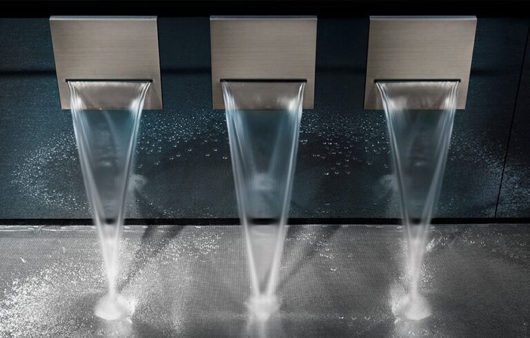 Three modern waterfall faucets flow onto a textured surface, creating a sleek, symmetrical water feature against a dark background.