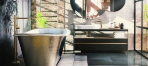 Modern bathroom with freestanding silver bathtub, brass fixtures, black vanity, round mirror, and large windows. Brick and leaf-patterned walls enhance elegance.