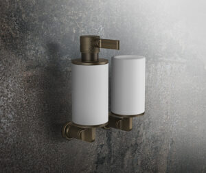 Two sleek, modern soap dispensers mounted on a textured, dark gray wall, featuring metallic bronze accents and minimalist design elements.