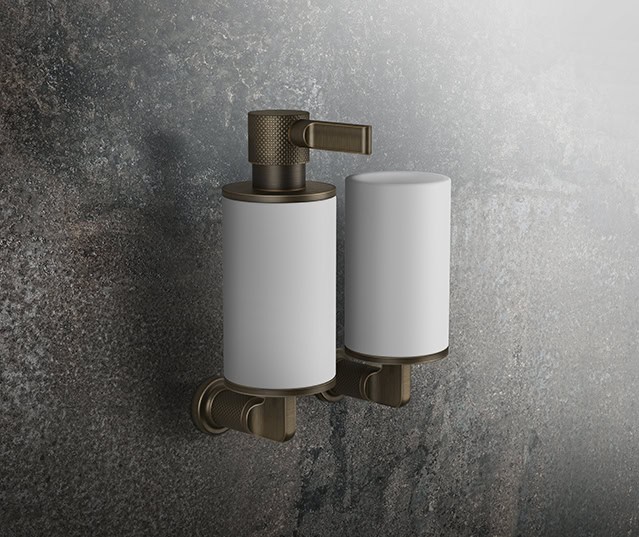 Two sleek, modern soap dispensers mounted on a textured, dark gray wall, featuring metallic bronze accents and minimalist design elements.