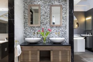 Elegant bathroom features dual sinks, mosaic tile backsplash, and a wooden vanity with fresh tulips. Modern lighting accents the sleek, stylish decor.