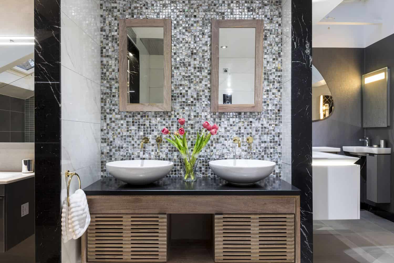 Elegant bathroom features dual sinks, mosaic tile backsplash, and a wooden vanity with fresh tulips. Modern lighting accents the sleek, stylish decor.