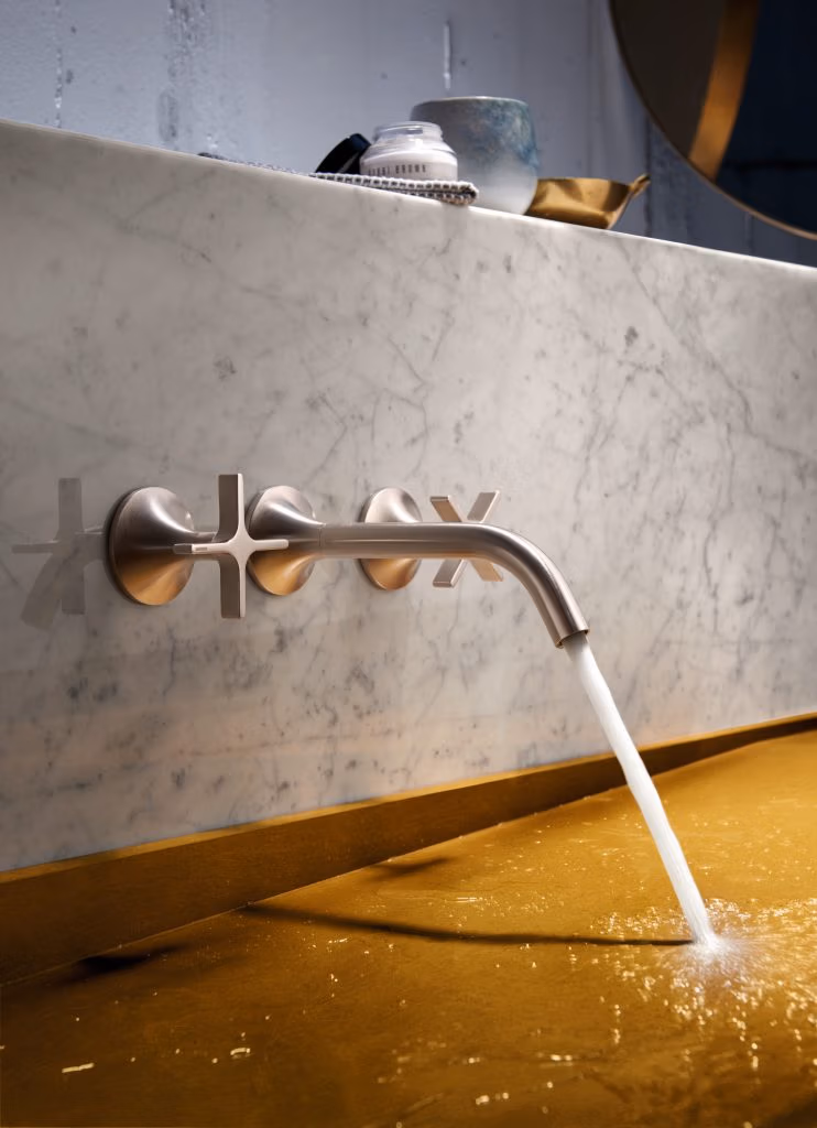 A modern stainless steel faucet with cross handles on a marble backsplash, pouring water into a golden sink. A cup and toiletries are seen on the counter.