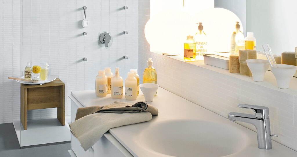 A modern bathroom with a countertop full of various soap bottles, a towel, sink, mirror, and neatly arranged toiletries on a wooden stool.