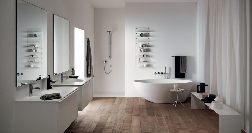 Modern bathroom with hardwood floors, dual sinks, large mirrors, free-standing bathtub, and minimalist decor. White walls and shelves create a clean aesthetic.