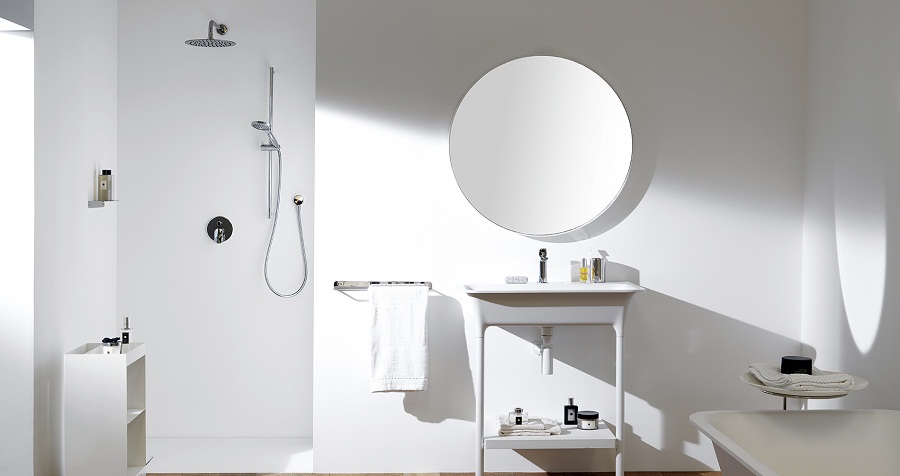 Minimalist white bathroom with round mirror, open shower, and simple sink. Shelving holds toiletries; a neatly folded towel hangs nearby.
