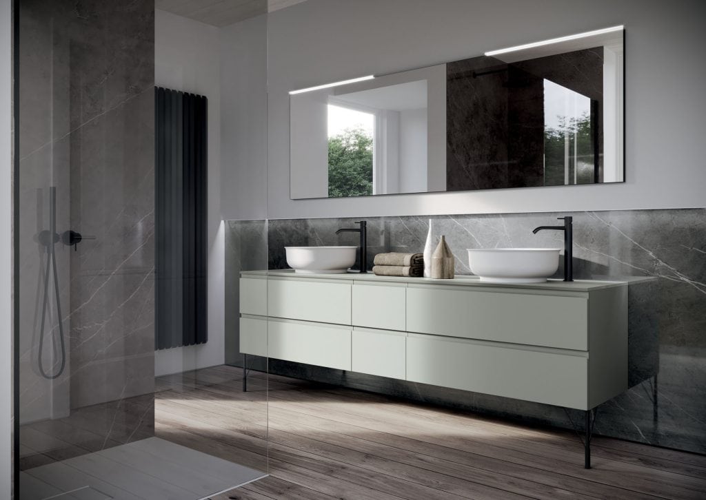 Modern bathroom with large mirror, sleek double sinks, and green cabinetry. Marble shower area, wooden floor, and window providing natural light complete the design.