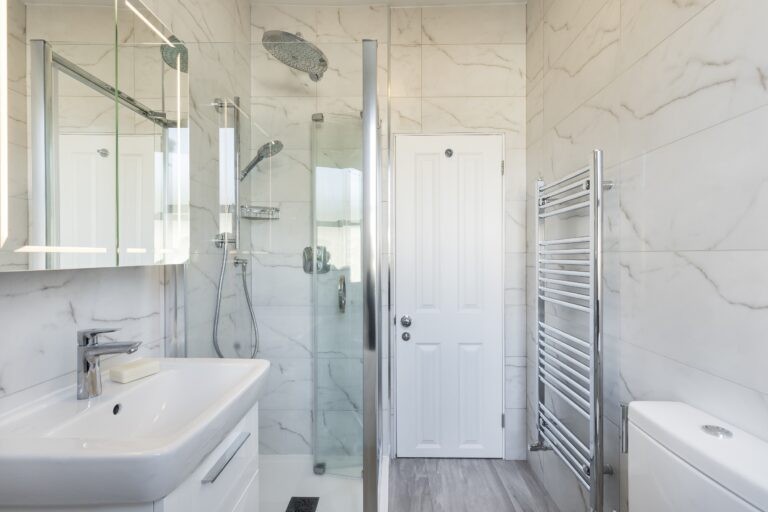 Modern bathroom with marble walls, sleek shower enclosure, white sink, wall mirror, and stainless steel towel rack. Bright, clean, and minimalistic design.