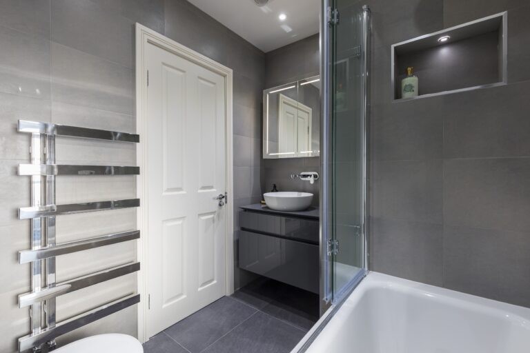 Modern bathroom with gray tiles, white door, glass shower screen, sleek basin, and towel rack. Spotlit recessed shelf holds a decorative bottle.
