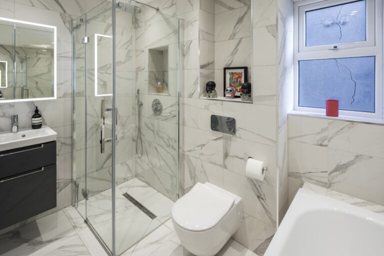 Modern bathroom with marble tiles, glass-enclosed shower, sleek fixtures, and a wall-mounted toilet. Minimal decor includes colorful artwork and small sculptures.