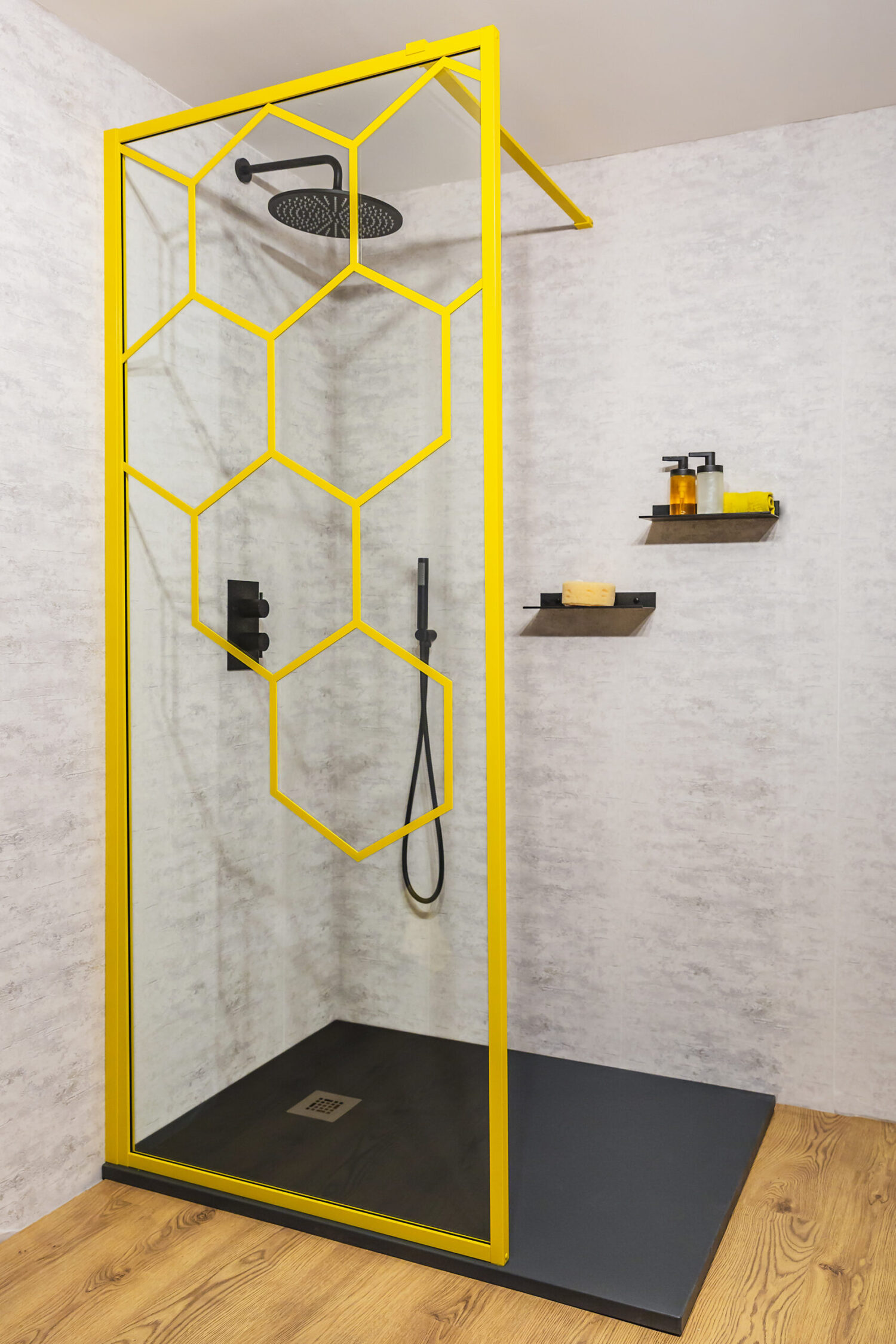 Modern shower with a yellow hexagonal frame, minimalist design. Grey walls, wooden floor, and shelves with bath products enhance the contemporary aesthetic.