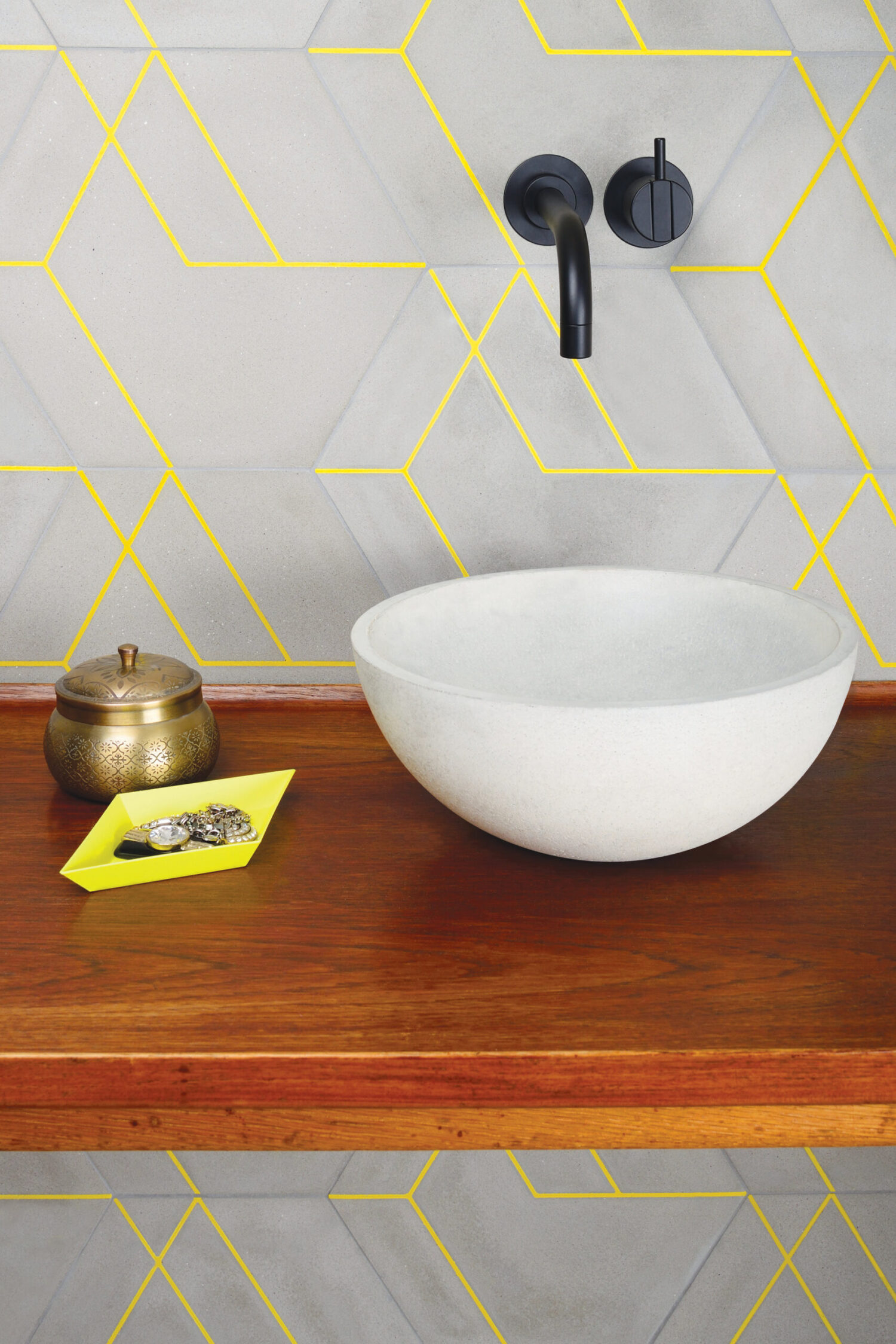 A stylish bathroom features geometric wall tiles, a round white sink on a wooden countertop, and decorative items in gold and yellow tones.