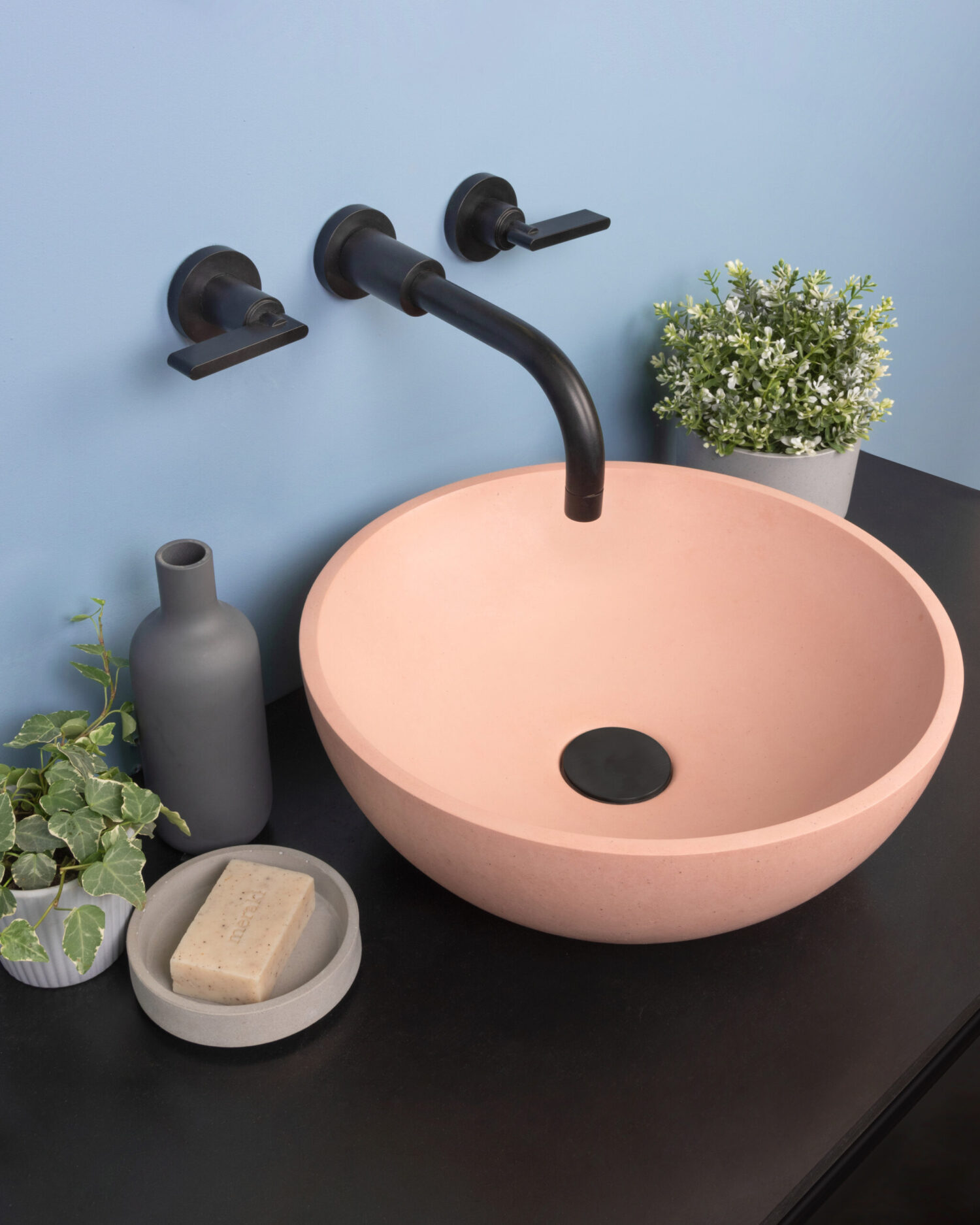 A modern bathroom sink setup with a pink basin, black faucet, soap, gray vase, and green plants, set against a pastel blue wall.