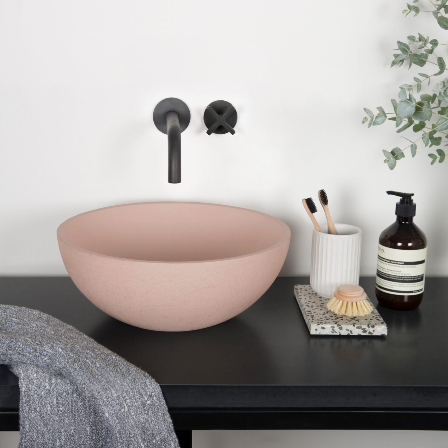 Minimalist bathroom scene with a pink vessel sink, black faucet, toiletries, and eucalyptus branches on a black counter, evoking a modern aesthetic.