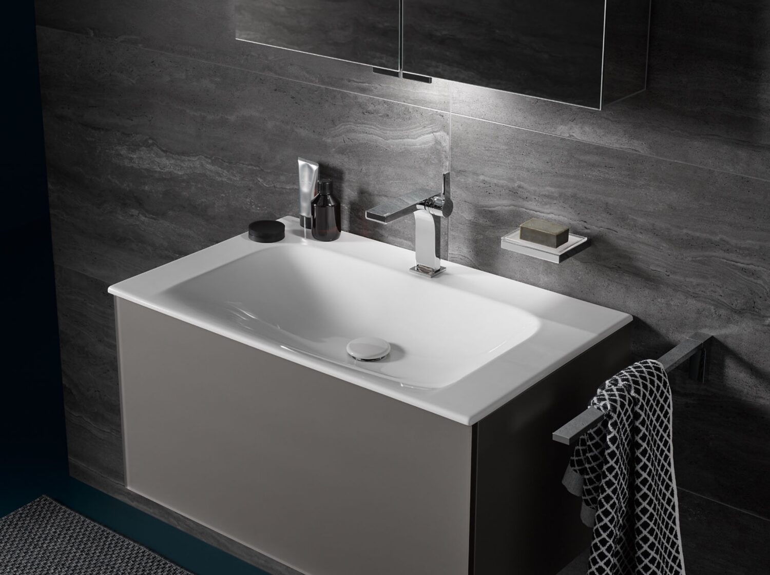 Modern bathroom with a white sink, chrome faucet, grey tile wall, and a towel hanging on a rail, featuring minimalistic design elements.