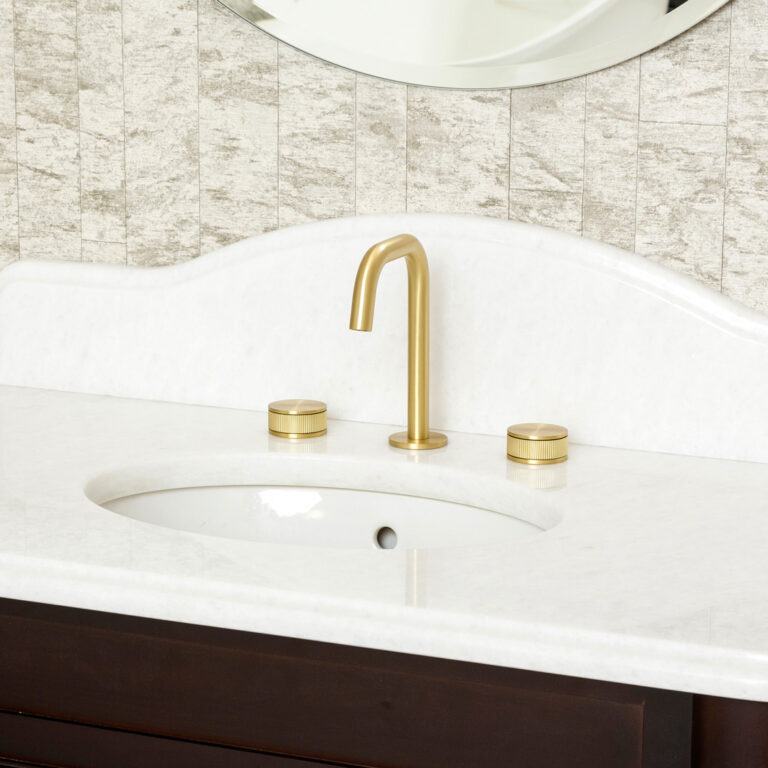 A modern bathroom vanity featuring a sleek, gold faucet with two matching handles on a white countertop, against a textured wall background.