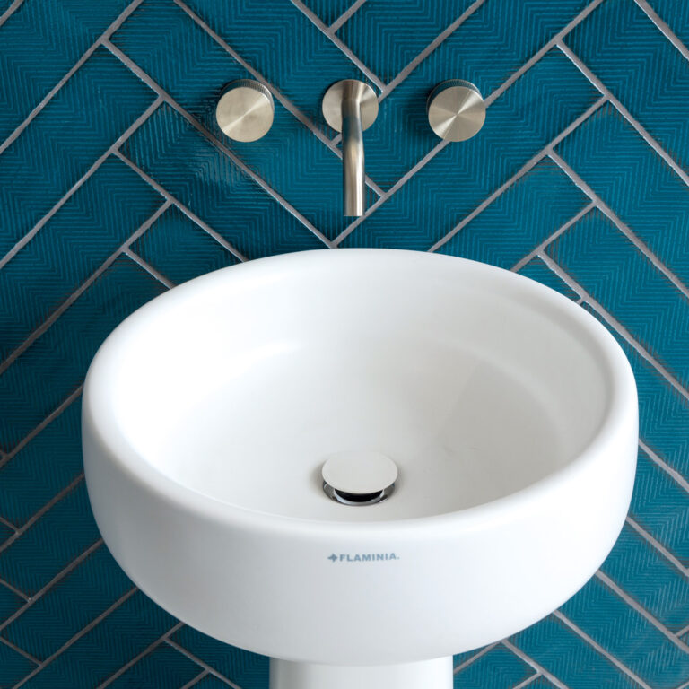 A white pedestal sink with stainless steel faucet and handles, set against a blue herringbone tiled wall, conveys modern bathroom design.
