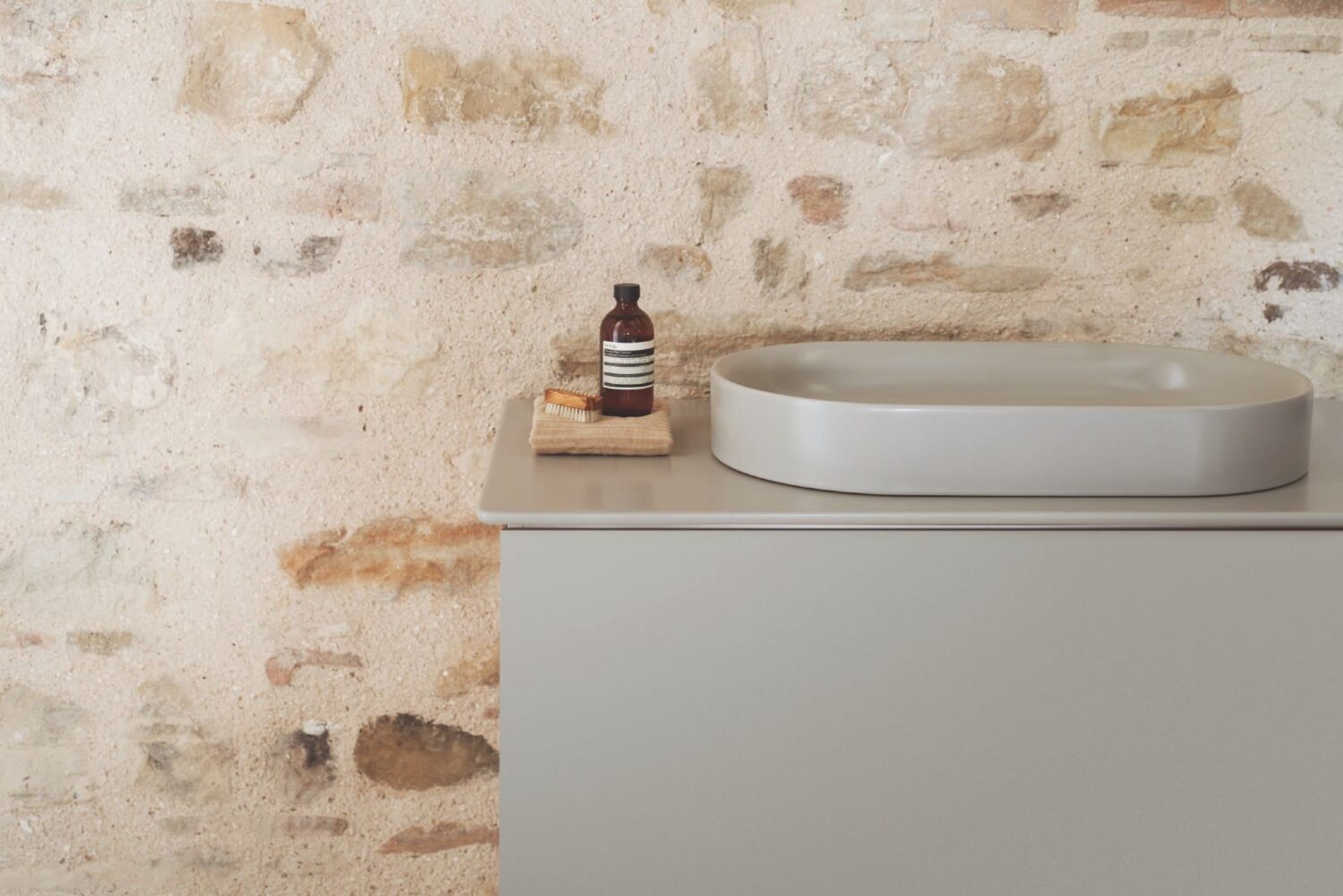 A minimalist bathroom sink with a bottle and brush on a textured stone wall background. Neutral tones create a calming aesthetic.