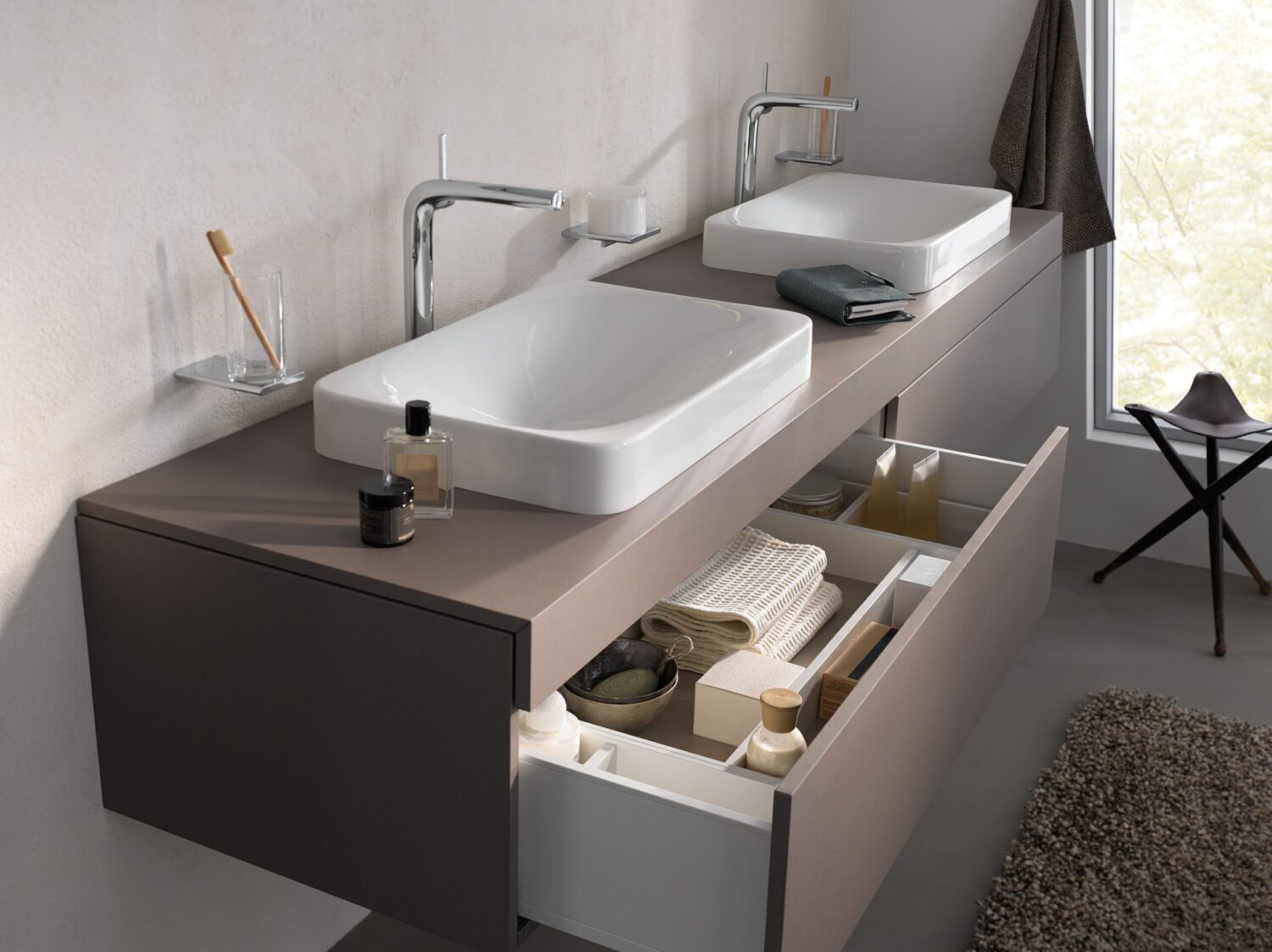 Modern bathroom with dual sinks, open drawers revealing towels and products. Minimalist decor includes a hanging towel and stool by the window.