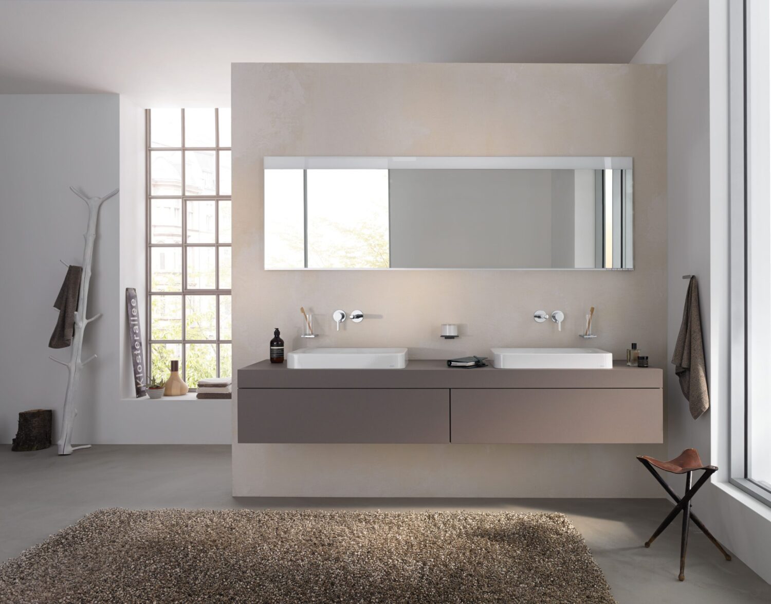 Modern bathroom with dual sinks, large mirror, and minimalist decor. Natural light from tall window, soft tones, and textured rug enhance elegance.