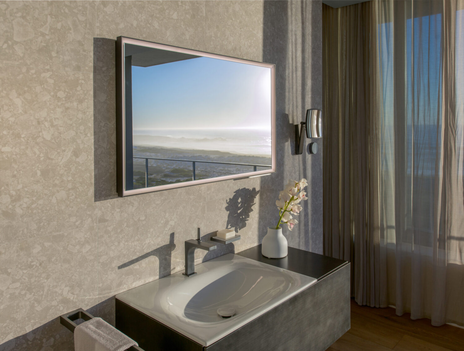 Modern bathroom with a sleek sink, large mirror reflecting an ocean view, elegant orchid vase, and light streaming through sheer curtains.