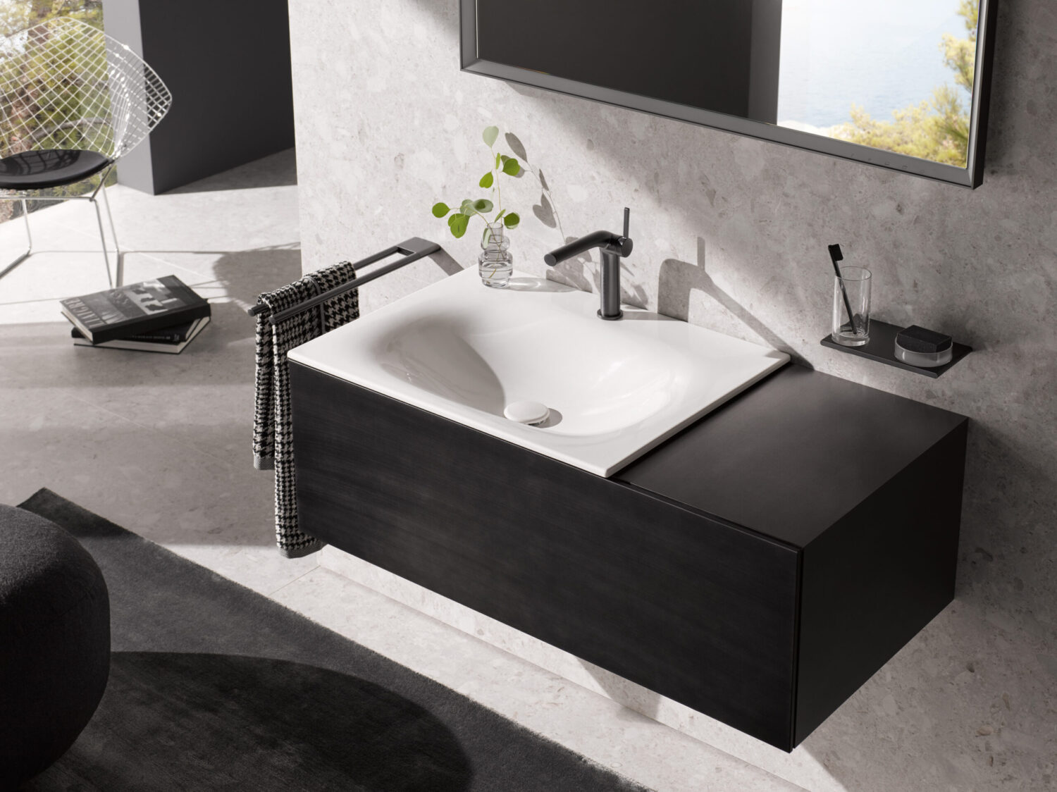 Modern bathroom with a sleek black sink, large mirror, and minimalist decor. A plant adds greenery beside a wire chair and books.