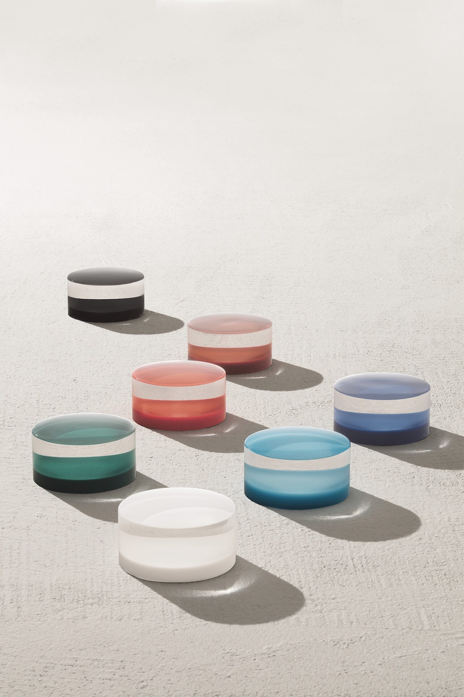 Seven colored, translucent round objects with white bands are arranged on a textured surface casting soft shadows in bright light.