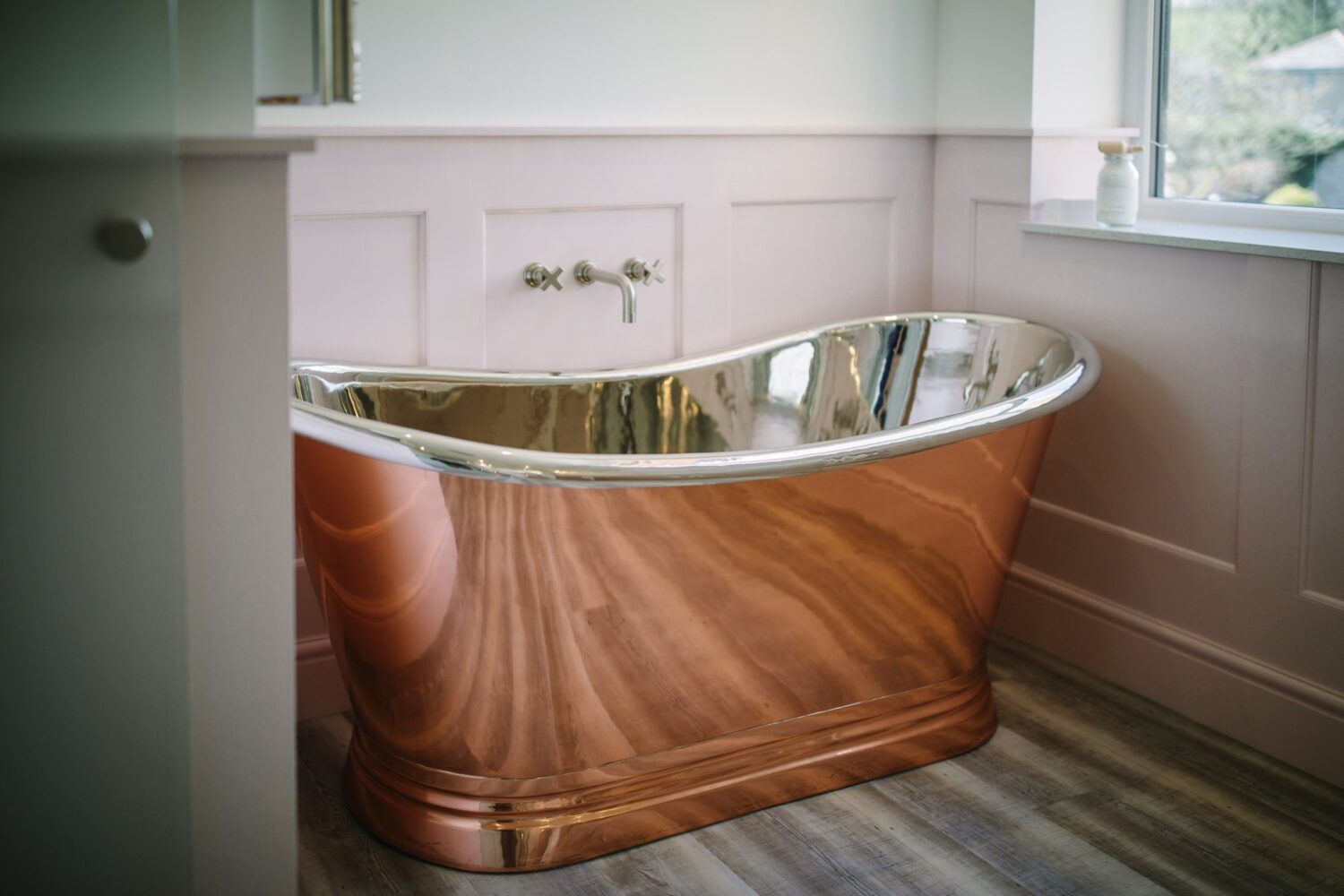 A luxurious copper bathtub in a stylish bathroom with wooden flooring and panelled walls, positioned near a window with natural light.