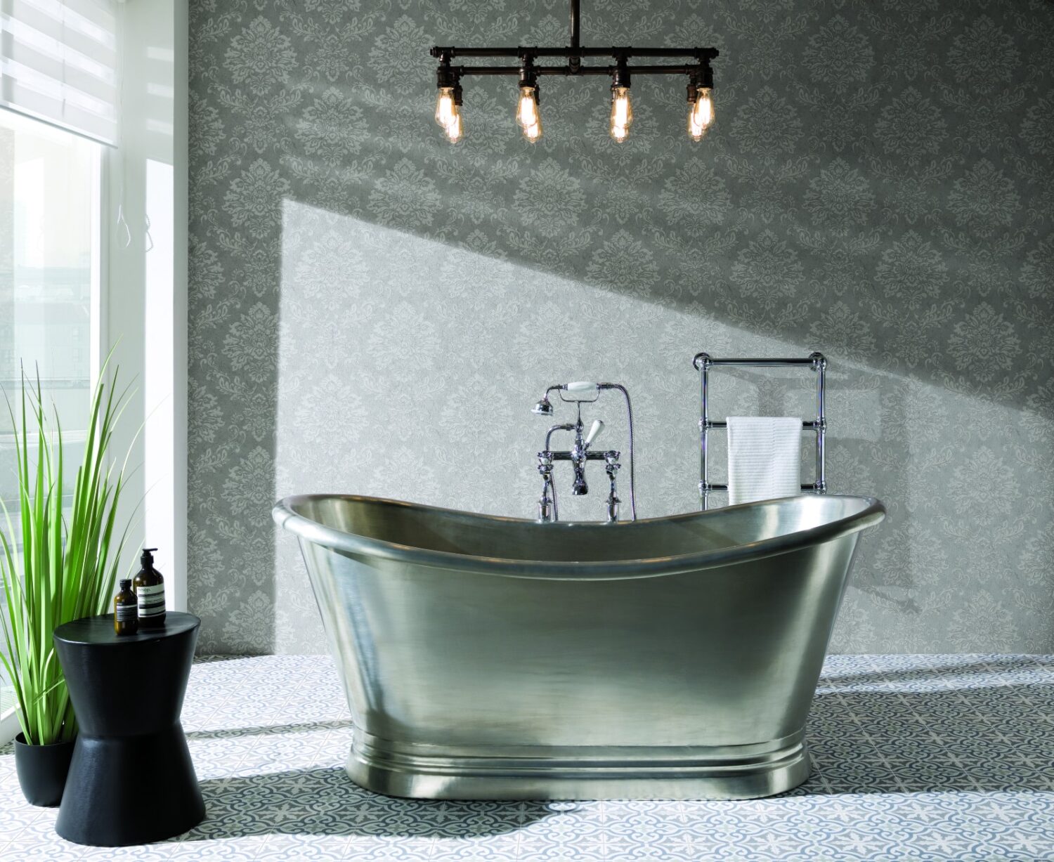 Luxurious bathroom features a metallic freestanding bathtub, elegant patterned wallpaper, industrial light fixture, potted plant, and side table with toiletry bottles.