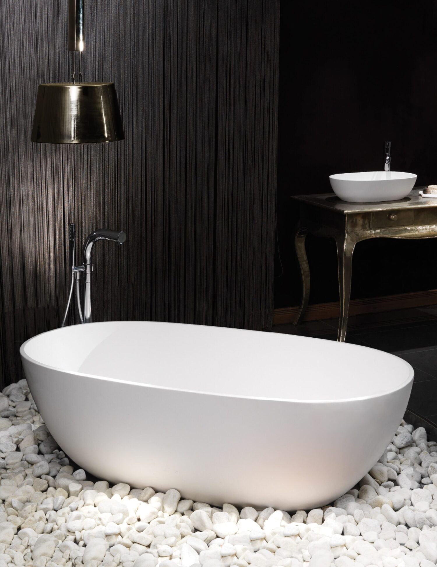 Modern bathroom with a sleek, white bathtub on white pebbles, metallic sink and lamp, set against a textured dark backdrop.