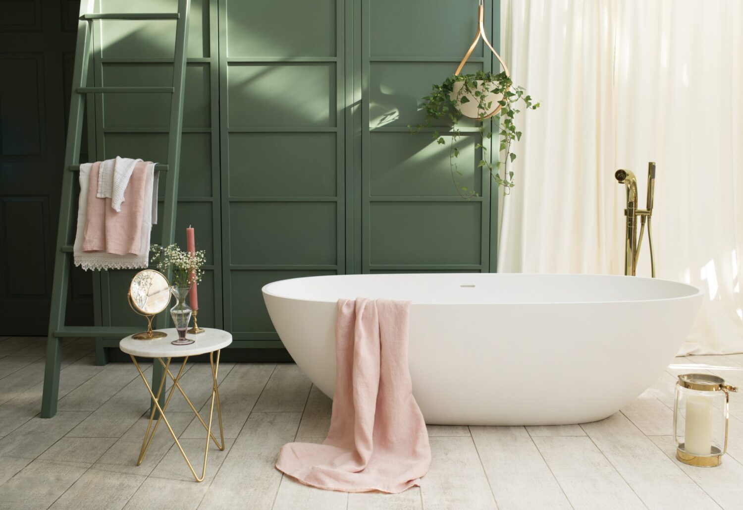 Bathroom with green paneled wall, white freestanding bathtub, pink towel, hanging plant, and small table with candle and mirror. Cozy, elegant atmosphere.