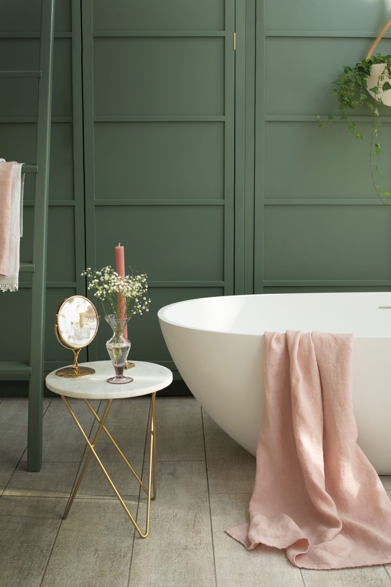 A stylish bathroom with a white tub, pink towel, elegant side table, candle, decorative plant, and a mirror against green panel walls.