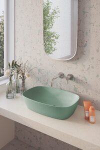 A modern bathroom features a green sink on a terrazzo counter, a wall mirror reflecting greenery, and minimalist decoration with small plants and bottles.