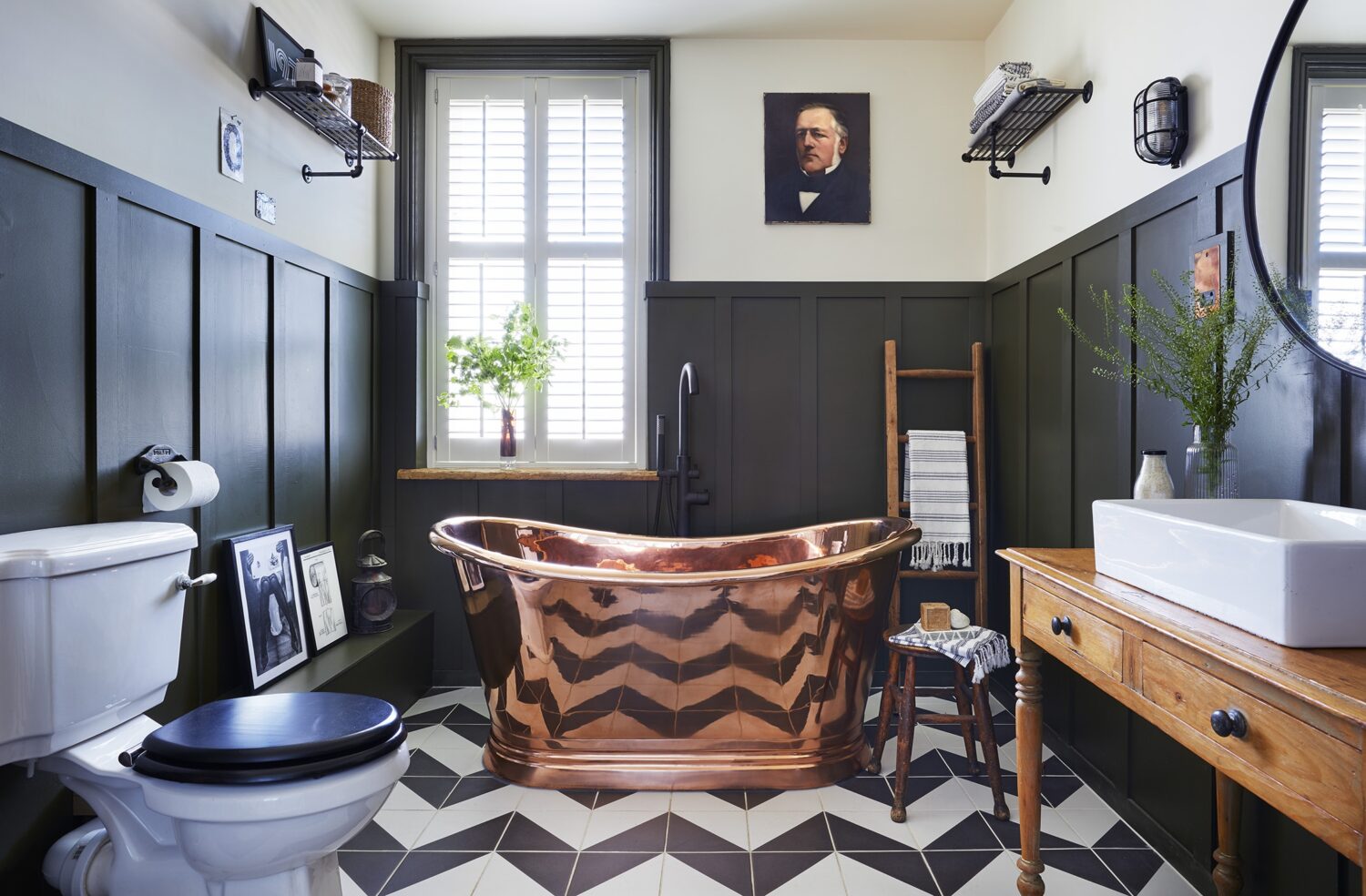 A stylish bathroom features a copper bathtub, vintage portrait, wooden accents, and geometric floor tiles beneath shuttered window and black paneled walls.