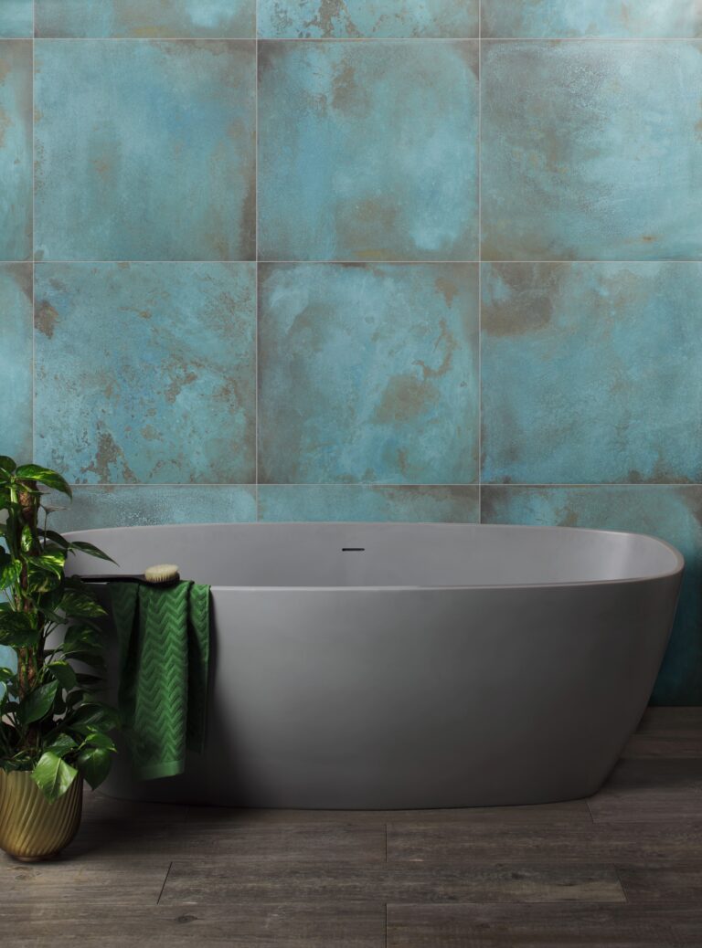 Modern bathroom with a sleek gray bathtub, green plant, and teal textured wall tiles. Wooden floor and green towel enhance the serene atmosphere.