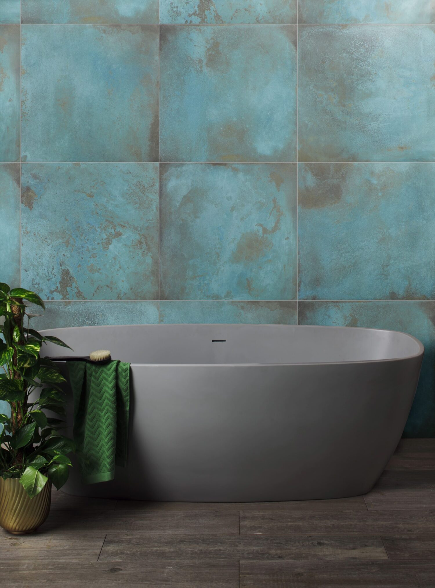 Modern bathroom with a sleek gray bathtub, green plant, and teal textured wall tiles. Wooden floor and green towel enhance the serene atmosphere.