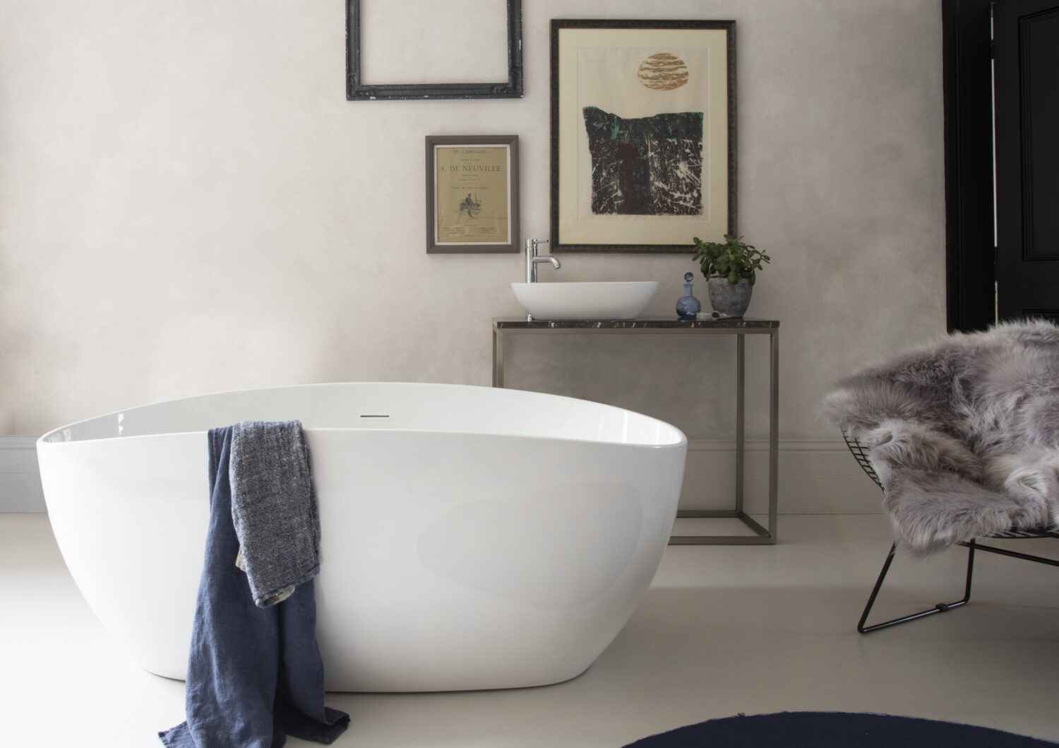 Modern bathroom featuring a sleek white bathtub, fluffy chair, minimal artwork, and a console with a vessel sink and decorative plant.