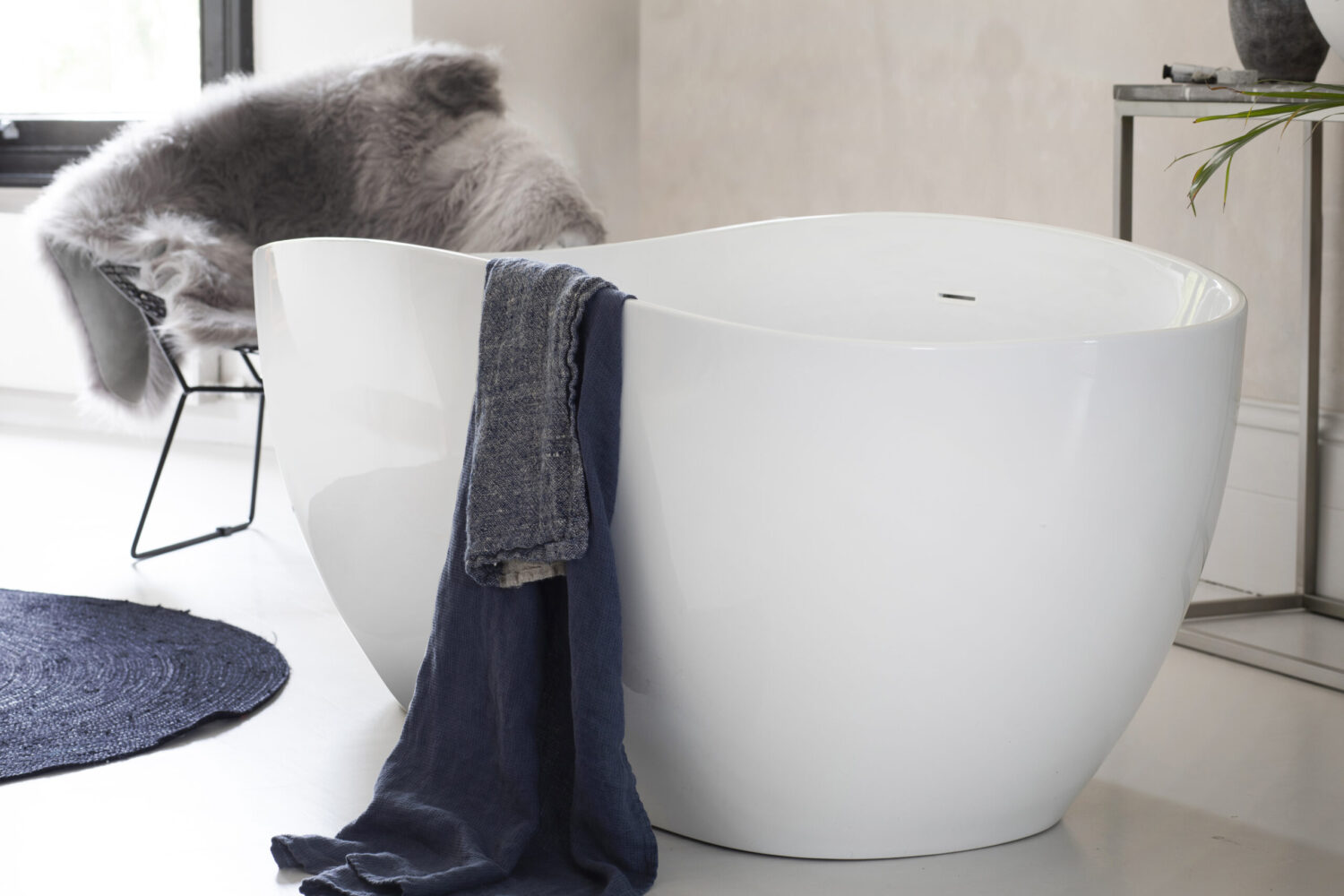 Modern bathroom features a sleek, white freestanding bathtub with dark towels. A fur-covered chair and a small table complete the minimalist decor.