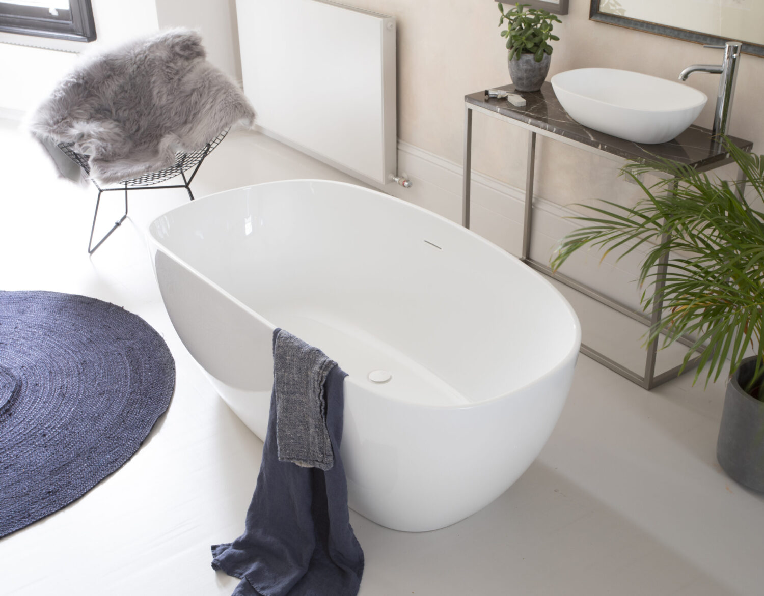 Modern bathroom with an oval tub, plant, and chair with fur throw. A dark blue rug accentuates the minimalist decor.