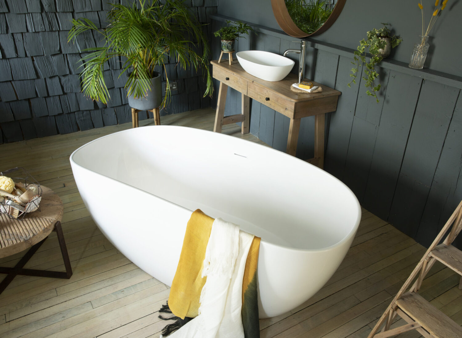 A modern bathroom features a freestanding bathtub, wooden vanity with vessel sink, round mirror, and plants, creating a serene, natural aesthetic.