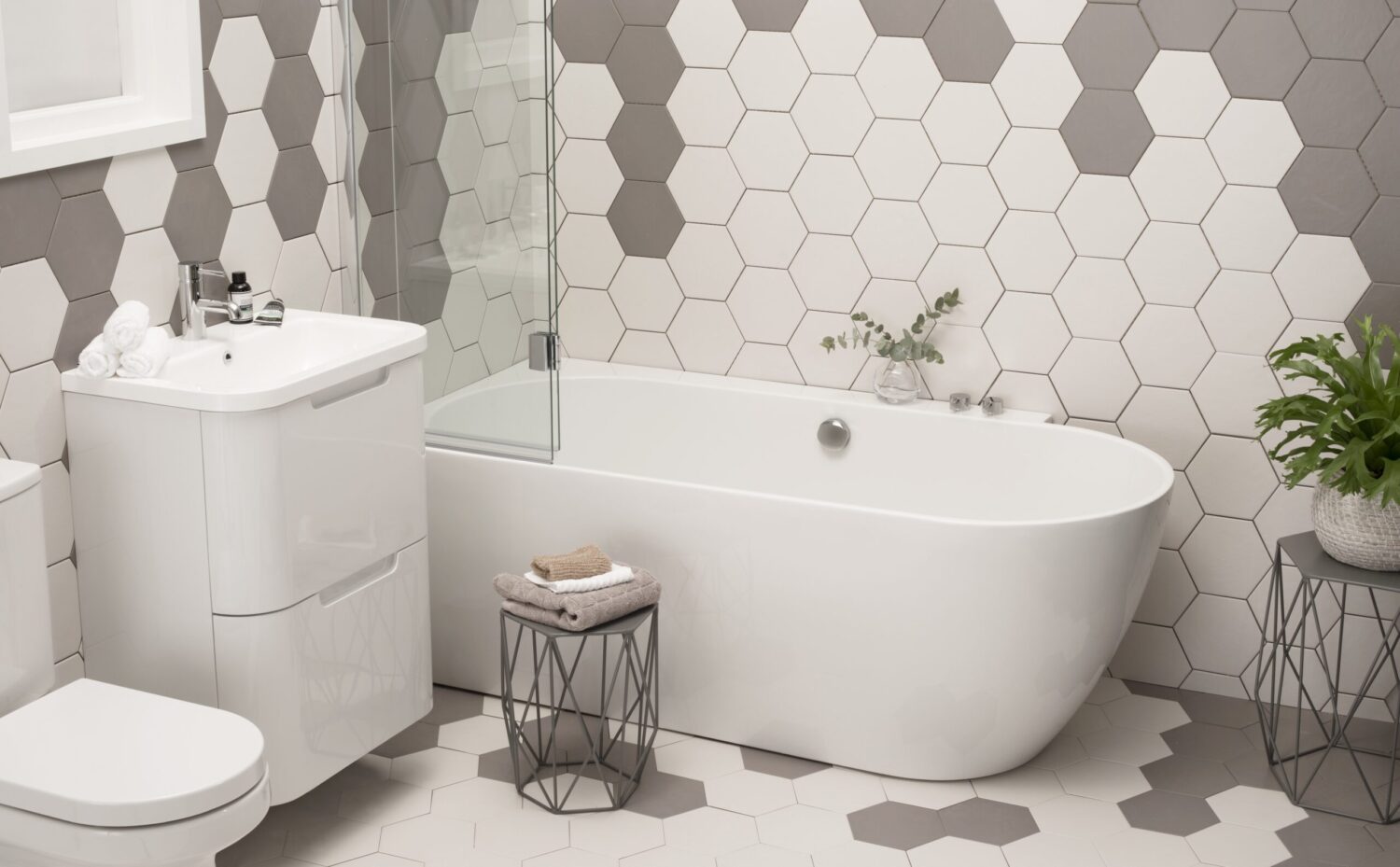 Modern bathroom with hexagonal tile design, white freestanding bathtub, compact sink, glass shower screen, green plant, and decorative table with folded towels.