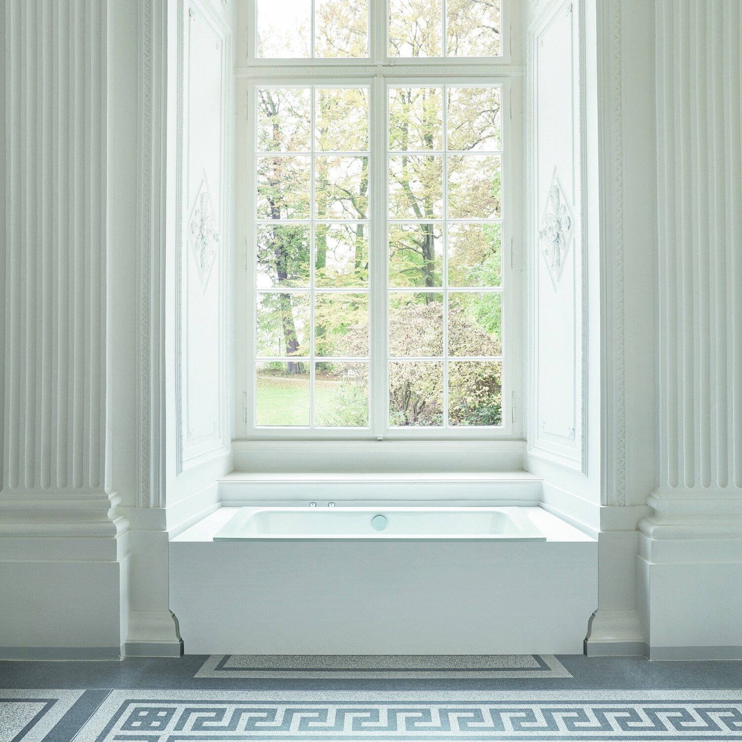 A luxurious white bathroom features a sunken bathtub, classical columns, and a large window overlooking a lush garden with autumn leaves.