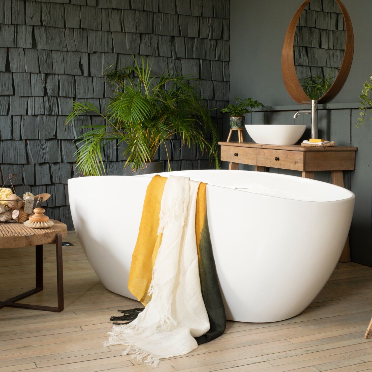 Modern bathroom with a freestanding bathtub, colorful towels, wooden vanity, potted plants, and a circular mirror. Warm, cozy atmosphere with natural accents.