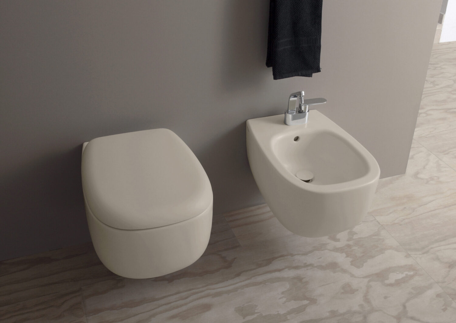 Modern bathroom with wall-mounted toilet and bidet on beige tiled floor, grey wall, and a black towel hanging above the fixtures.