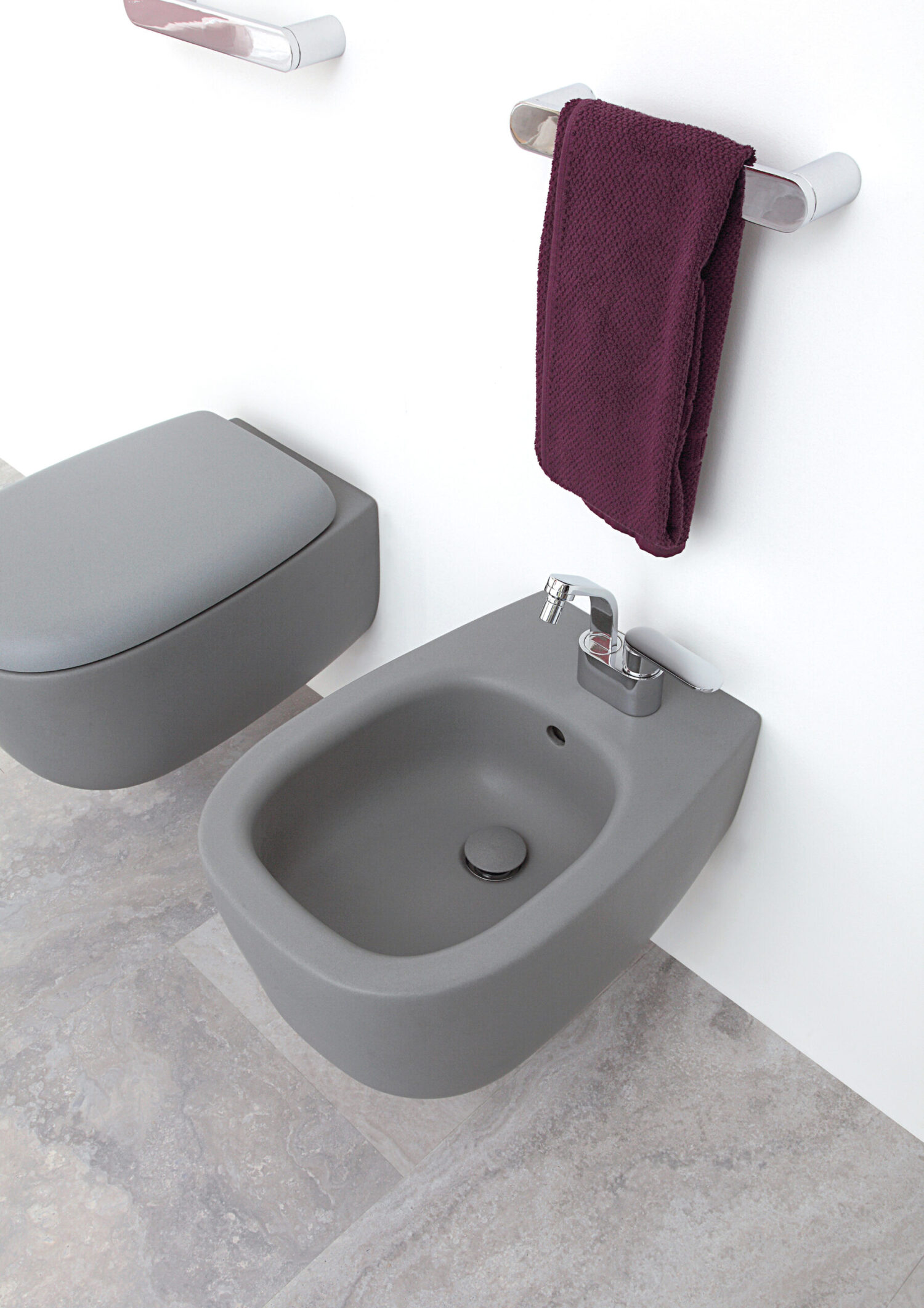 Gray bathroom with wall-mounted toilet and bidet. Dark purple towel hangs on the holder. Neutral stone tile floor enhances modern aesthetic.