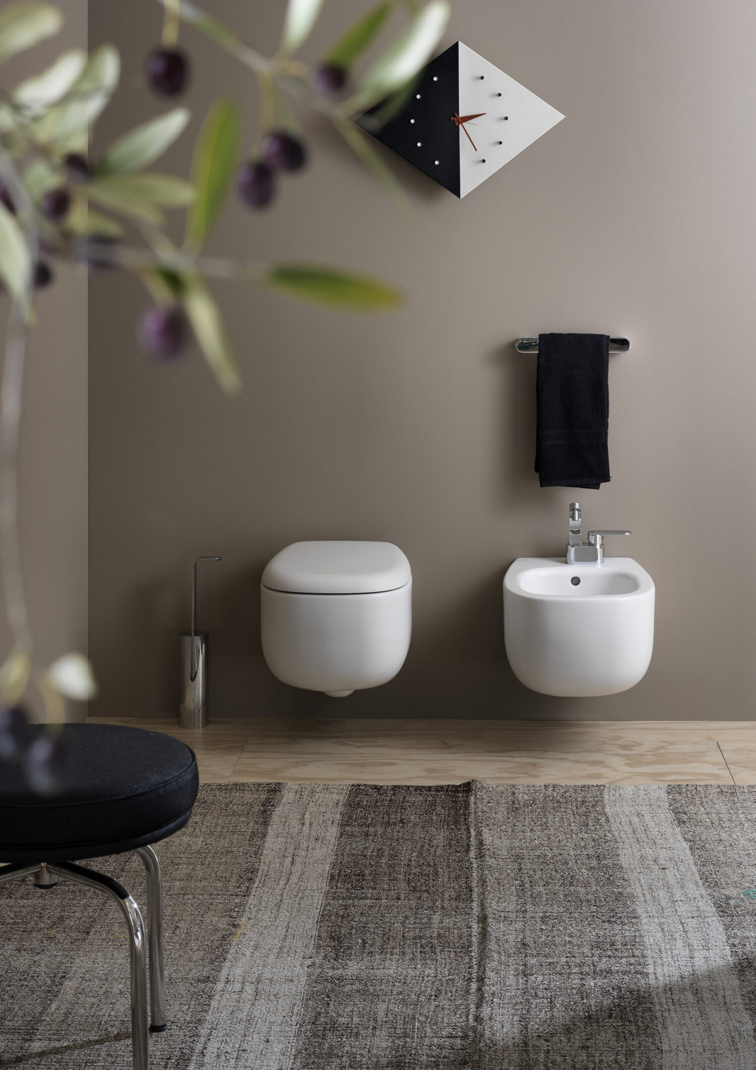 Modern bathroom interior with wall-mounted toilet and bidet, black towel, geometric wall clock, and olive plant in the foreground on a textured rug.