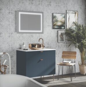 Modern bathroom with decorative tiles, illuminated mirror, and a gold sink. Wooden chair holds towels; green plant adds a natural touch.