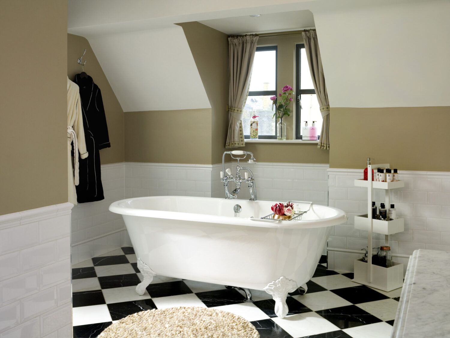 Elegant bathroom with clawfoot bathtub, checkered floor, and wall-mounted shelves. Robes hang beside a window with decorative curtains and flowers.