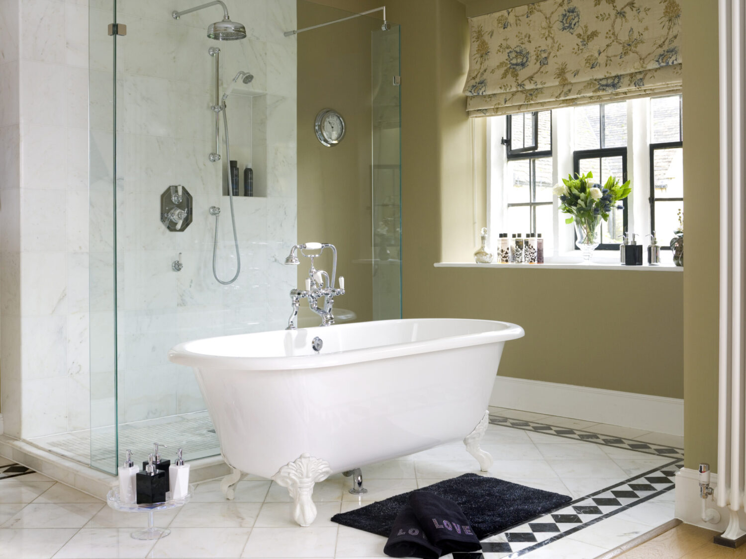 Elegant bathroom featuring a clawfoot tub, glass shower, marble tiles, and sunlit window with floral drapes. Decor includes plants and toiletries.