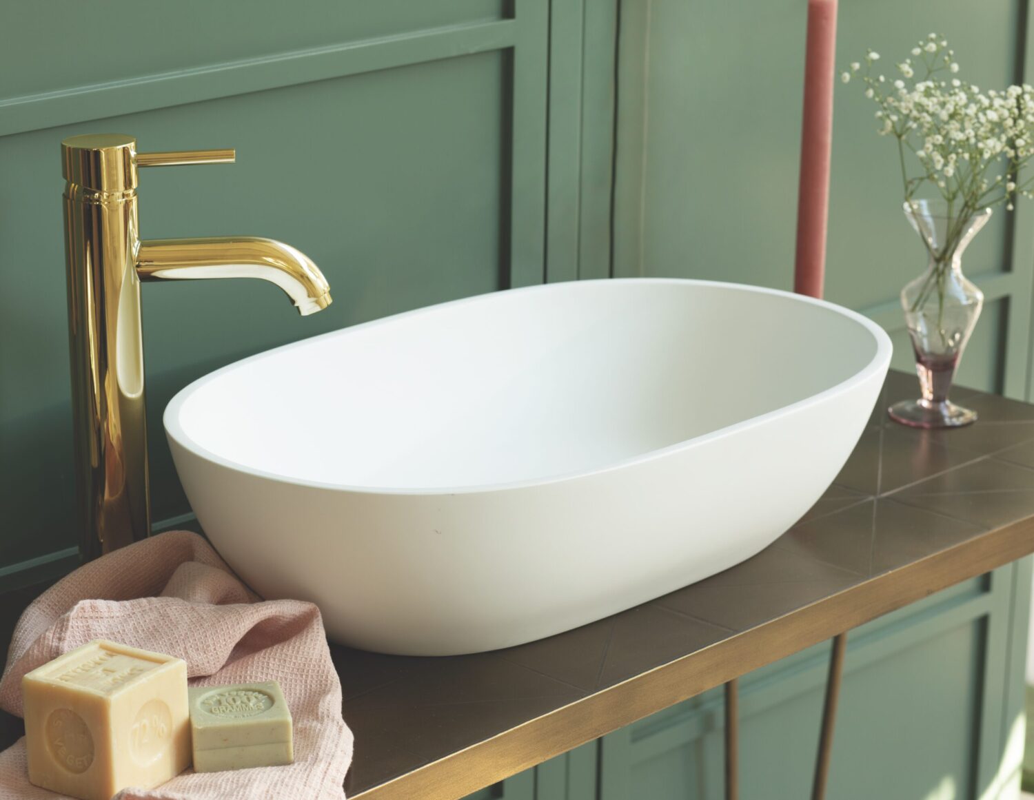 Elegant bathroom with a white oval basin, gold faucet, pink towel, and decorative soaps. Green paneling and a vase add style.